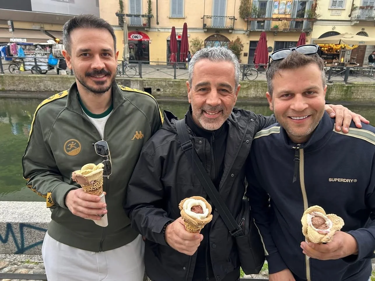 Three men smiling and holding ice cream cones, standing near a canal in a European city, with shops and buildings in the background.