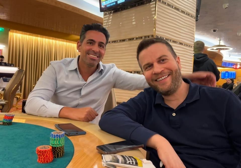 Two men smiling and sitting at a poker table in a casino, with poker chips, a mobile phone, and a newspaper in front of them.