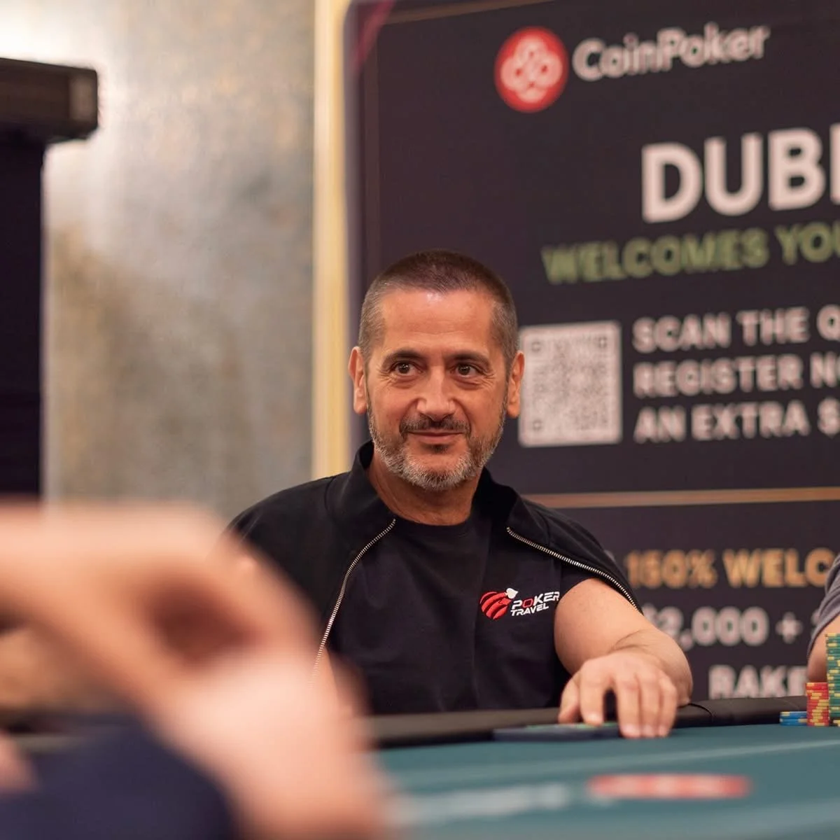 A man with short hair and a beard sitting at a poker table, with a PokerTravel shirt, in front of a dark sign with information about a poker event.
