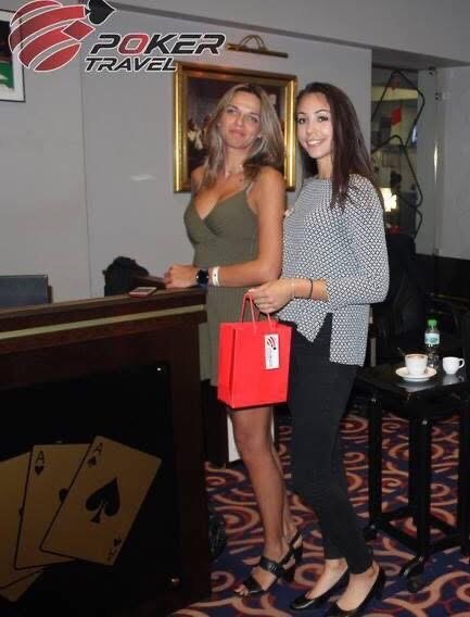 Two women standing at a poker tournament, one holding a red gift bag, in an indoor setting with poker-related decor and glasses on a table.