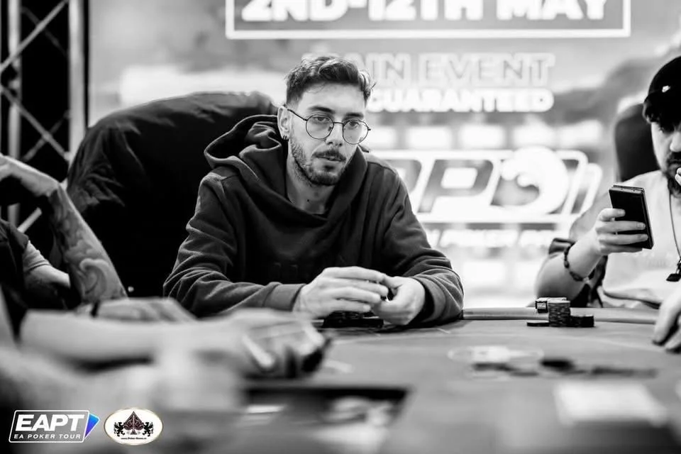 A man with glasses and a beard sitting at a poker table, holding poker chips, surrounded by other players, with a large promotional banner in the background.