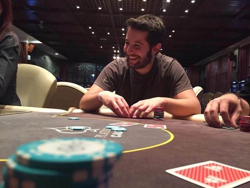 A man sitting at a poker table, smiling and playing cards with poker chips in front of him, in a casino or poker room setting.