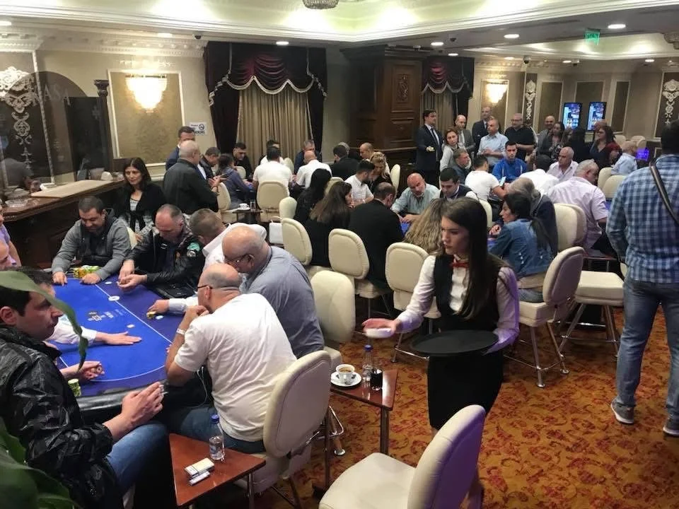 A crowded casino room with people gathered around poker tables playing poker, and waitresses serving drinks. The room has elegant decor with chandeliers and drapes.