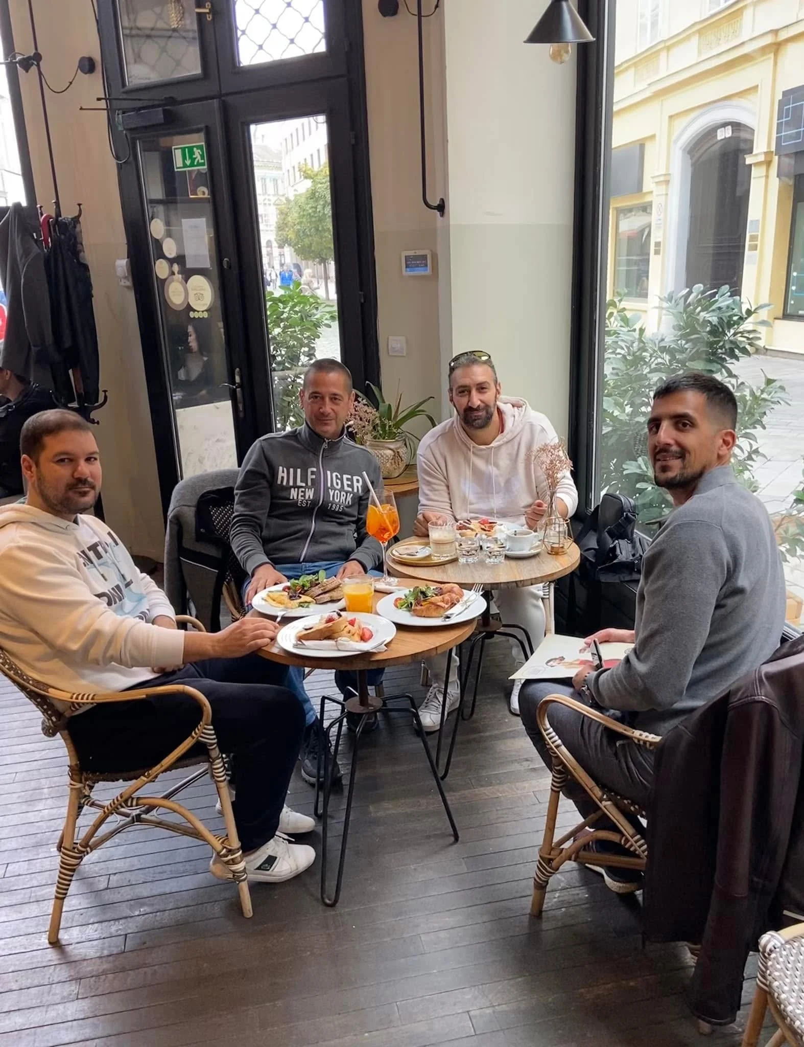 Five men sitting at a round wooden table in a café, enjoying a meal with plates of food, drinks, and coffee. The café has large windows with a view of a street outside. The men are smiling and dressed casually.