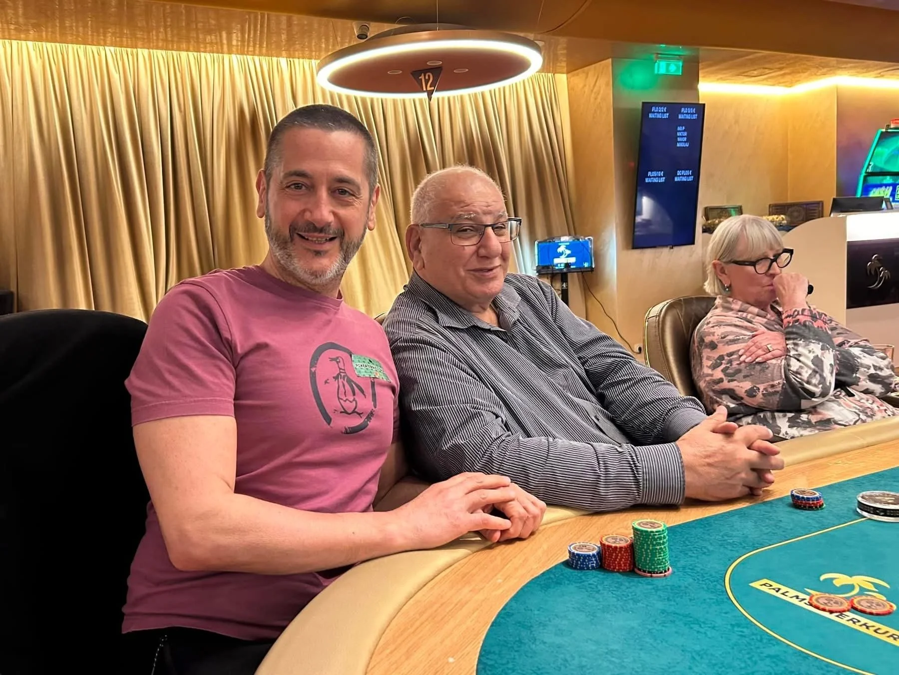 Three people sitting at a poker table with poker chips in front of them. The background has gold curtains and a large circular light fixture hanging from the ceiling. The man on the left is smiling and wearing a pink shirt, the man in the middle is w