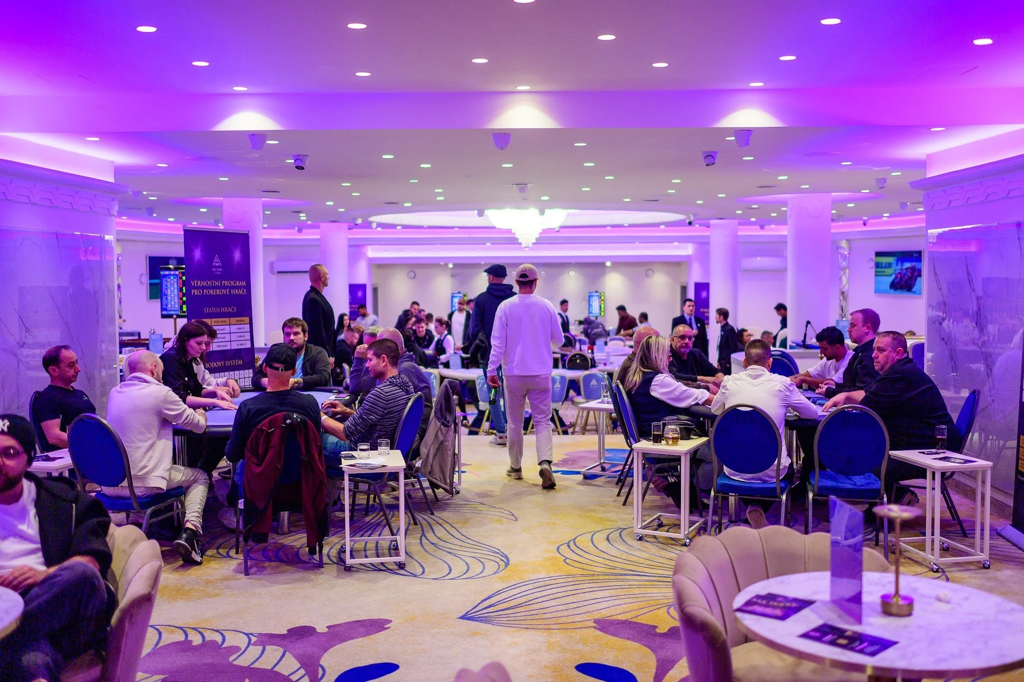 A large casino hall with purple and blue lighting, filled with people playing poker at numerous tables. Some individuals are engaged in conversation, while others are focused on their game. The room features elegant chandeliers, patterned carpet, and