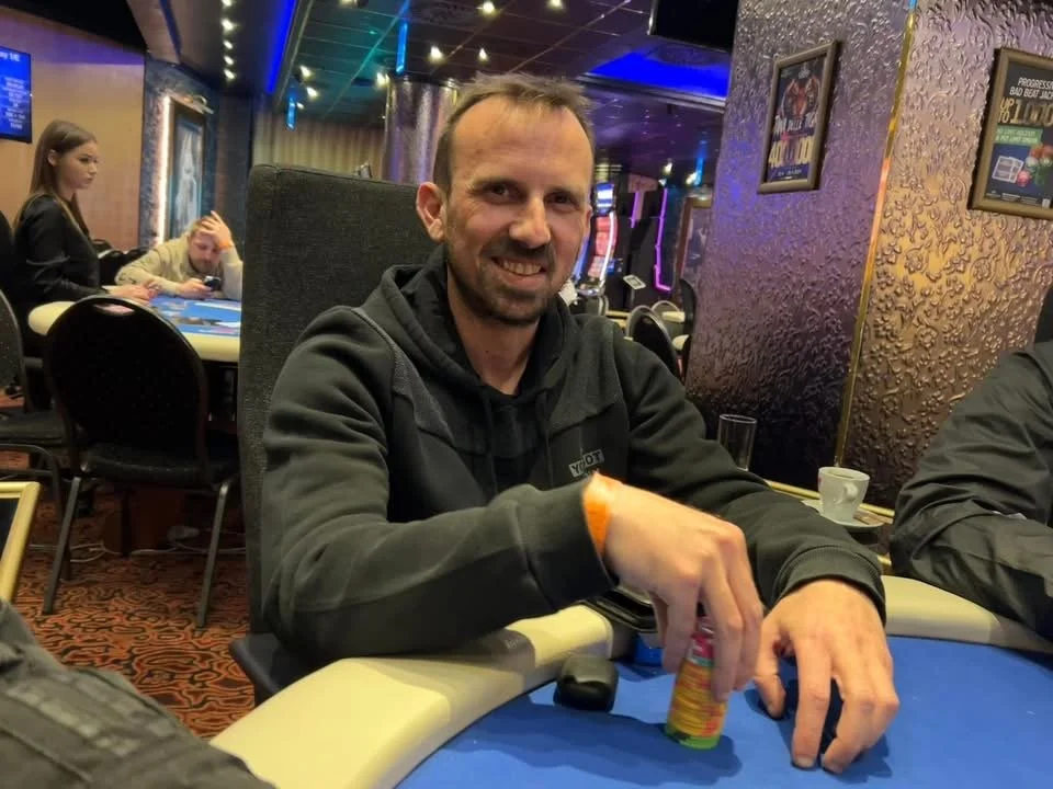 A man with a beard and short hair smiling while stacking poker chips at a casino table. In the background, there are other people sitting at tables, some with playing cards, and slot machines are visible in a brightly lit gaming area.
