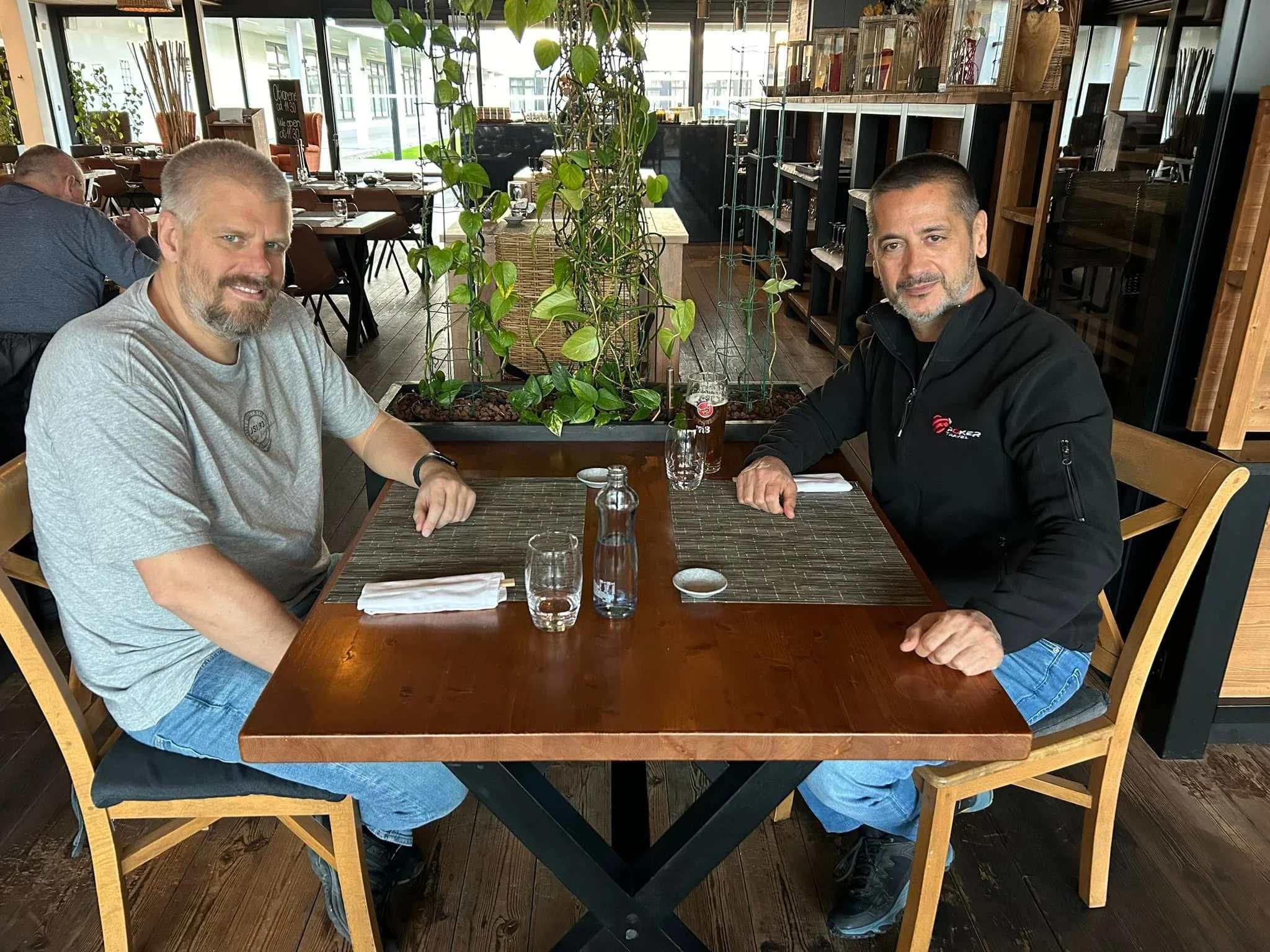 Two men sitting at a wooden table in a restaurant or cafe, shaking hands and smiling. The table has glasses, a bottle, and napkins. Behind them are large windows, green plants, and other patrons dining.