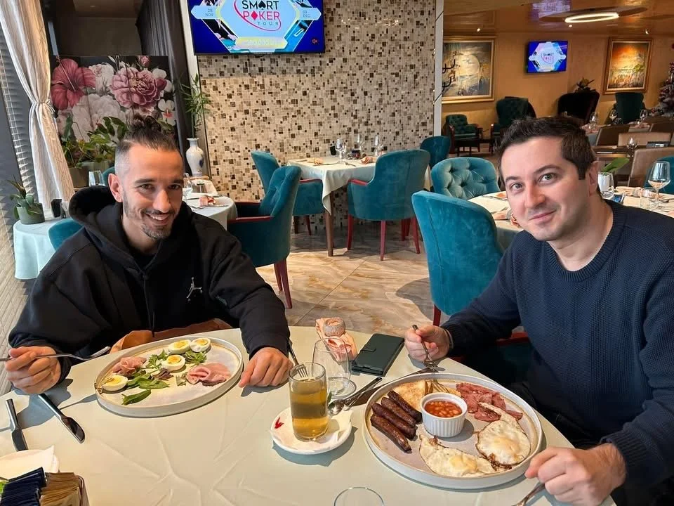 Two men sitting at a restaurant table with plates of breakfast food, including eggs, bacon, sausage, and a glass of beer. The restaurant has teal chairs and a decorative tile wall, with a large floral mural and multiple televisions on the walls.