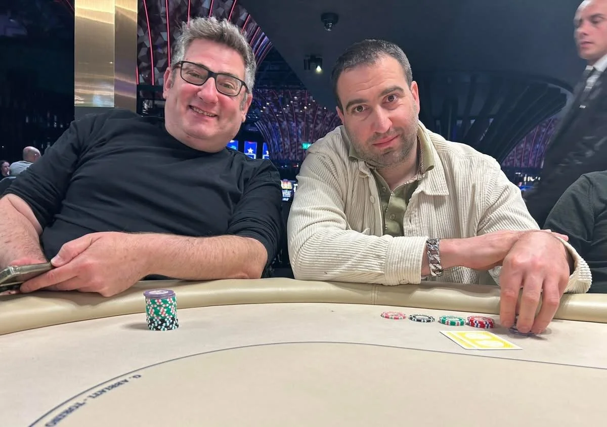 Two men at a poker table with poker chips and cards, smiling at the camera inside a casino.
