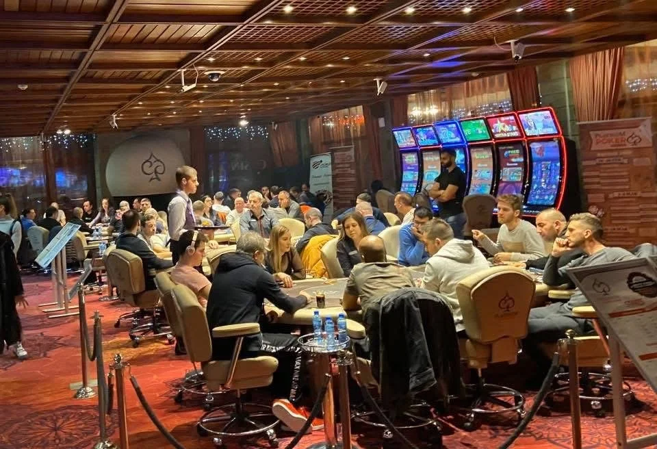 People playing poker at tables in a large casino or gaming room, with electronic gaming machines in the background. The room has a wooden ceiling and warm lighting.