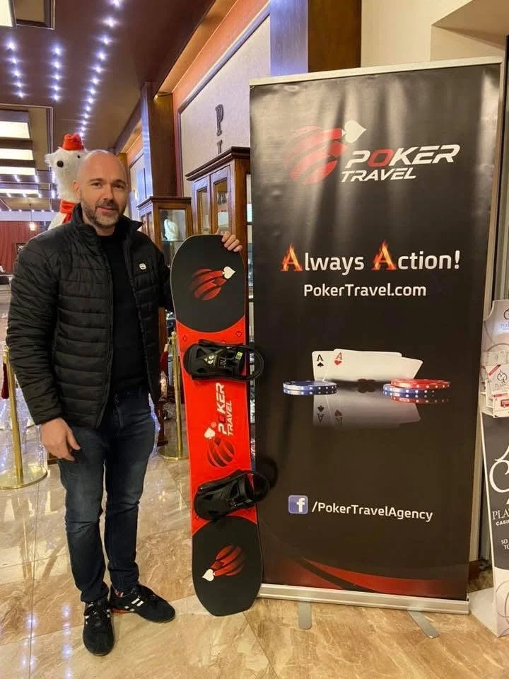 A man standing next to a snowboard with a PokerTravel logo and promotional banner for PokerTravel.com and Facebook page in an indoor setting.
