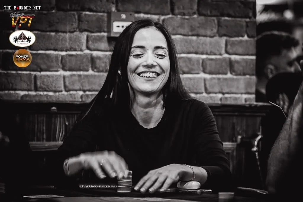 A woman smiling while playing poker at a table with poker chips, in a setting with an exposed brick wall.