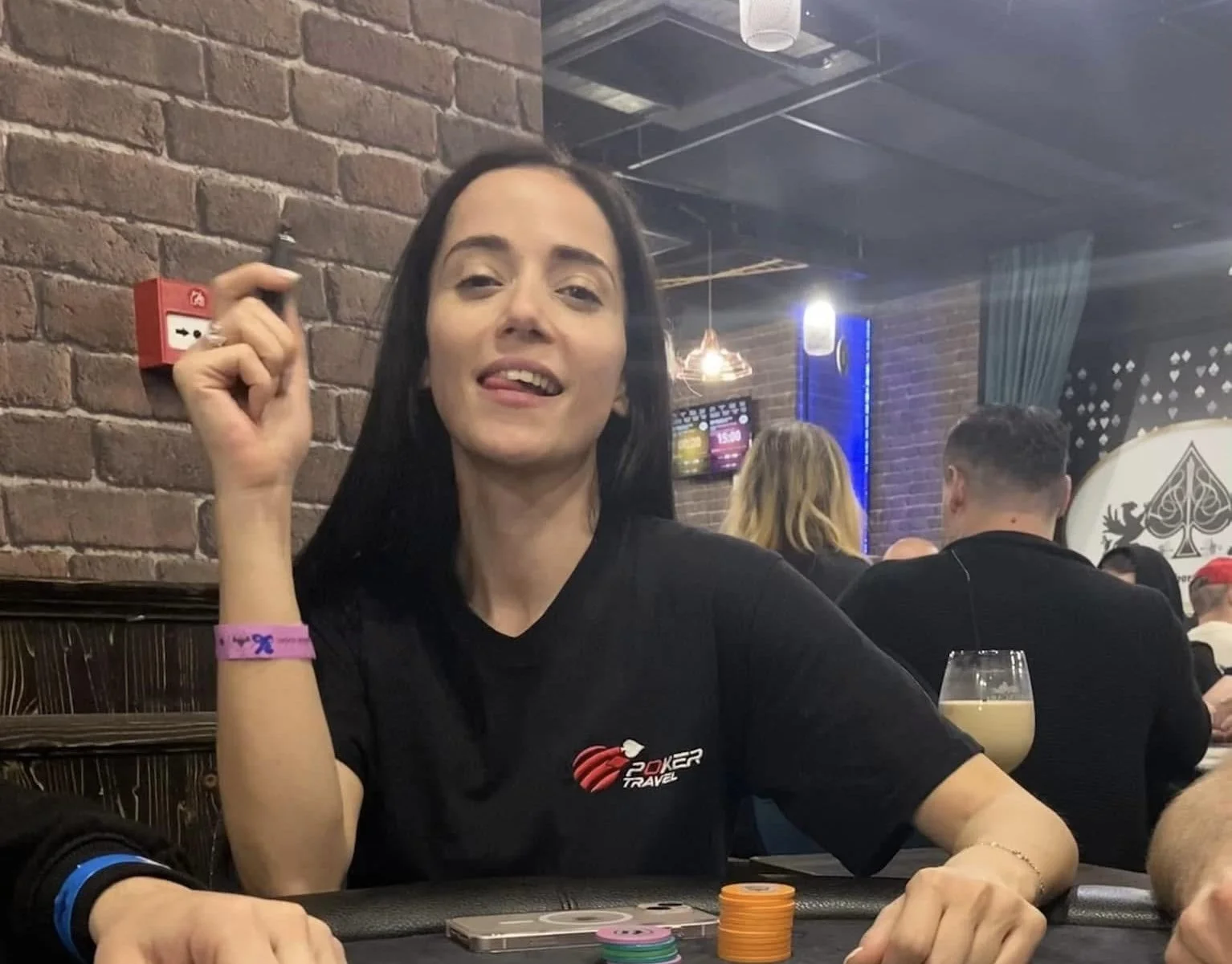 A woman with long dark hair sits at a poker table in a casino or gaming venue, holding a card in her right hand. She is wearing a black T-shirt with the Poker Travel logo, a pink wristband, and has poker chips in front of her. Other people are sittin