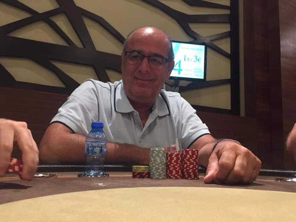 A man wearing glasses and a light gray polo shirt is sitting at a poker table with poker chips and a water bottle in front of him. He is smiling and looking at the camera. In the background, there is a screen displaying part of a poker tournament inf