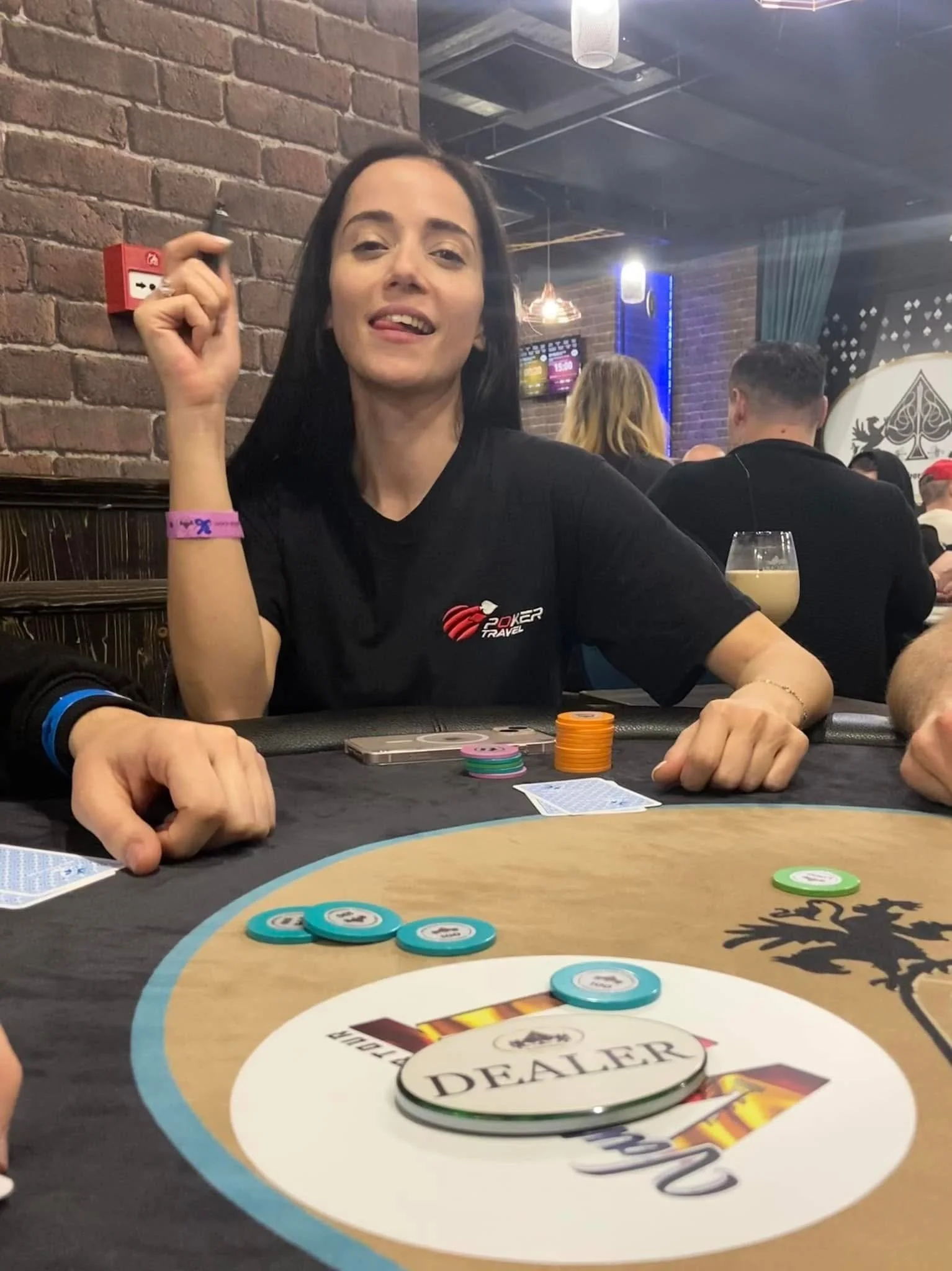 A woman with long dark hair, wearing a black shirt with a poker logo, sitting at a poker table with poker chips and cards, in a restaurant or bar setting with brick walls and pendant lighting, surrounded by other people playing poker.