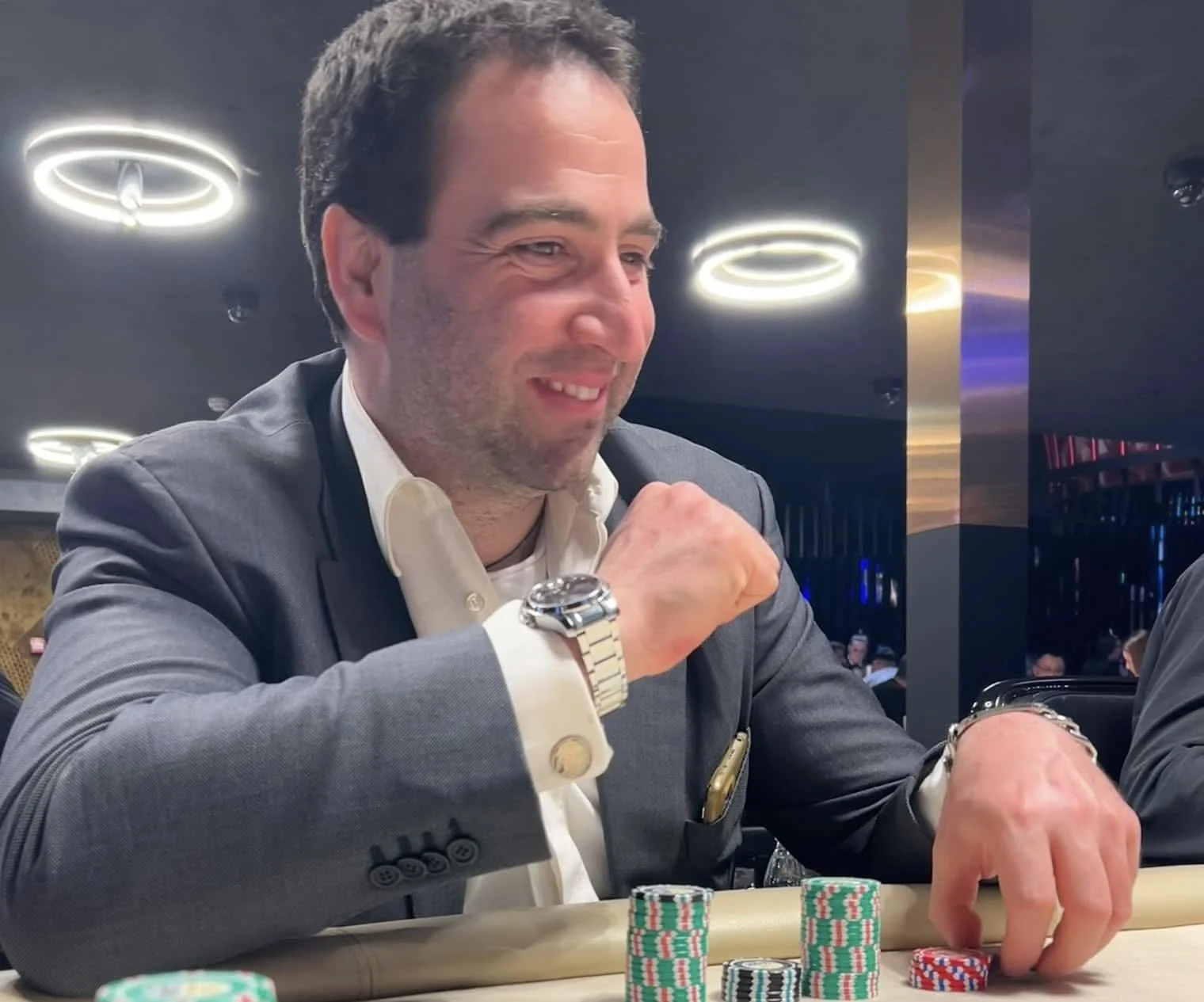 A man in a suit with a white shirt and a silver watch is playing poker at a casino table with poker chips in front of him, smiling and focused on the game.