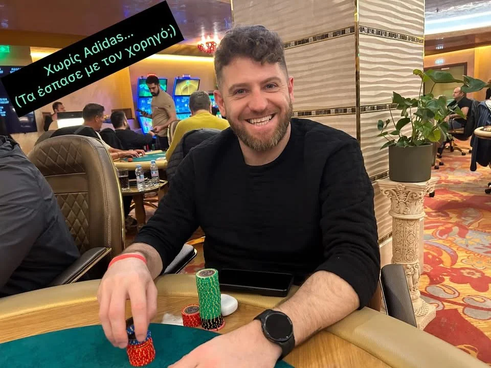 A man with a beard and curly hair smiling at a casino poker table, stacking poker chips. The background shows other people playing casino games and a partially visible slot machine.
