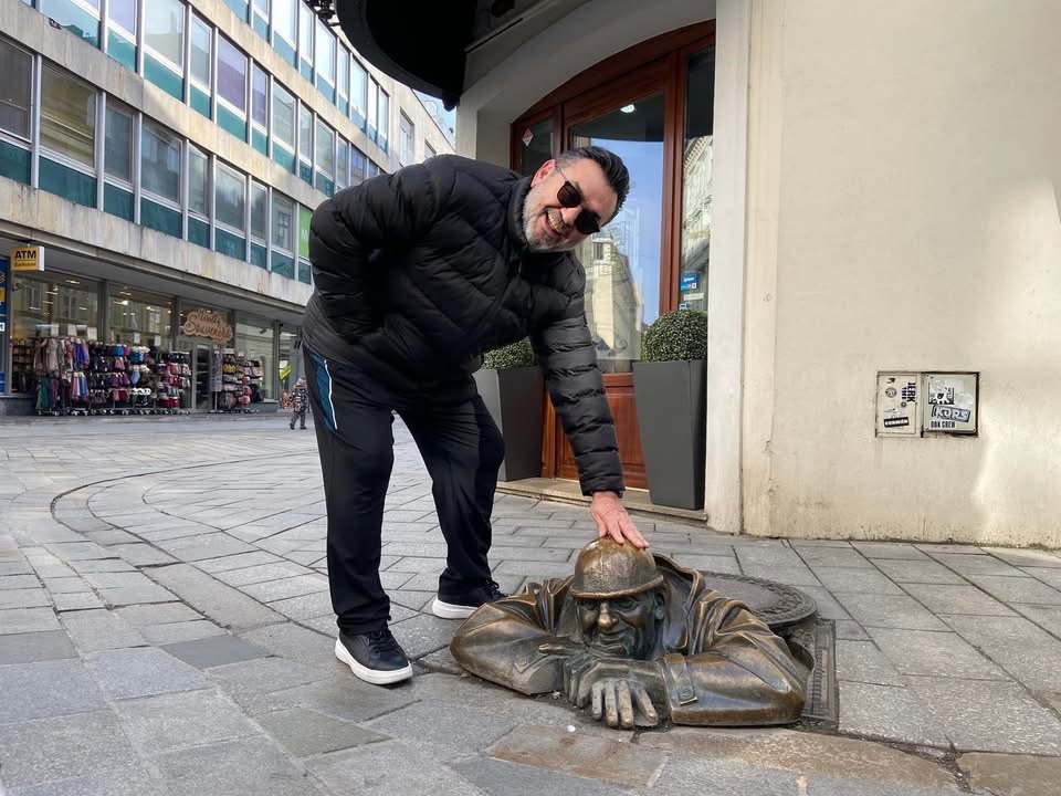 A man dressed in black with sunglasses crouching and touching the head of a bronze statue of a man with a hat, who appears to be emerging from the ground on a city sidewalk.