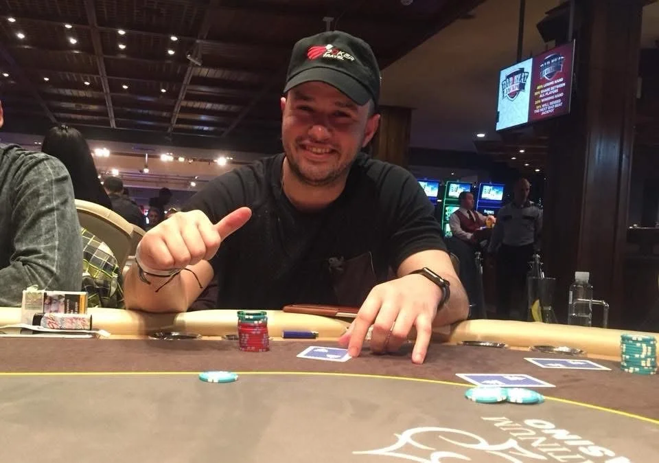 A smiling man in a black shirt and cap sitting at a poker table with poker chips and playing cards, giving a thumbs up in a lively casino or poker room.