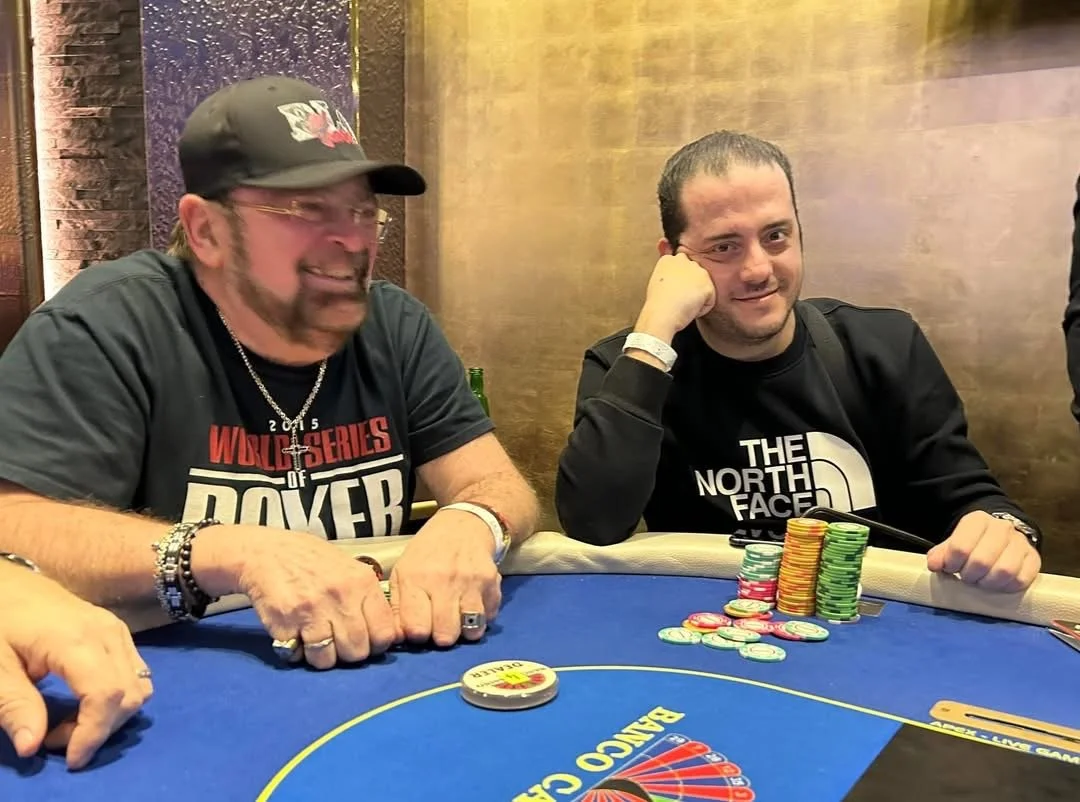 Two men sitting at a poker table with poker chips in front of them. The man on the left is wearing a black cap and a black t-shirt with red and white text, smiling. The man on the right is wearing a black North Face jacket, resting his head on his ha