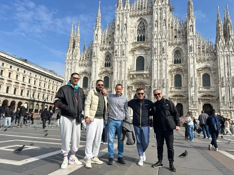 Five men standing in front of the Milan Cathedral in Italy, with people and pigeons around.