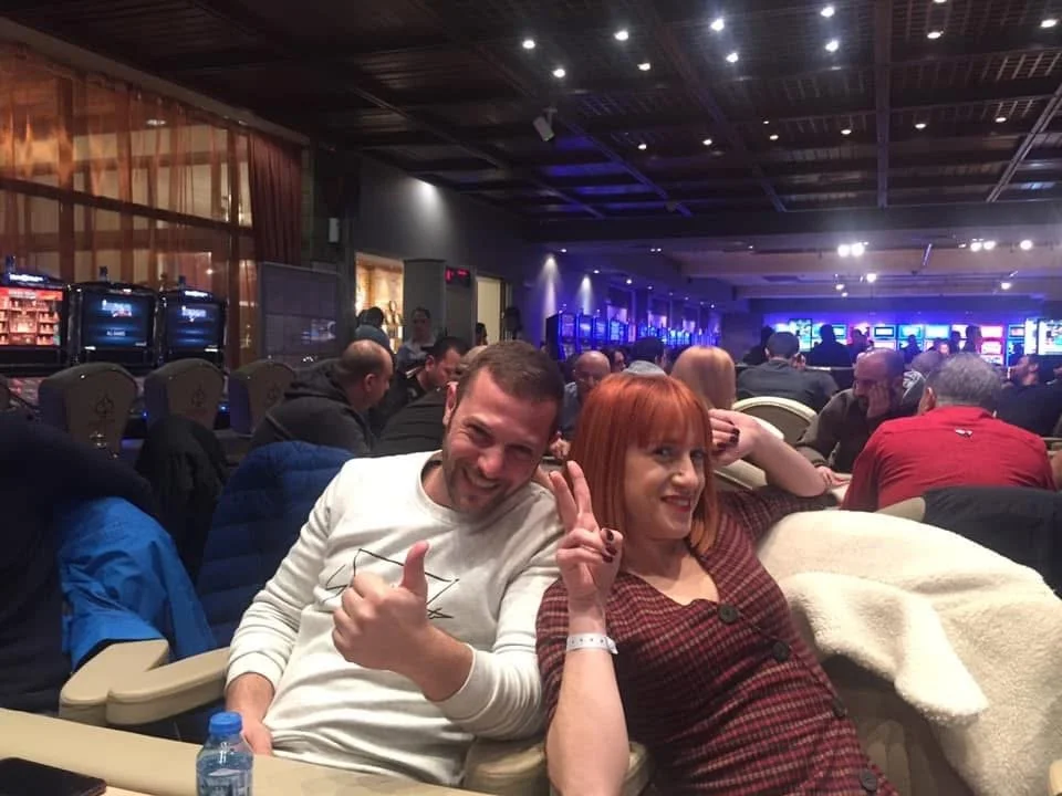 Two smiling people sitting in a crowded casino, with slot machines in the background, at a gaming event or casino night.