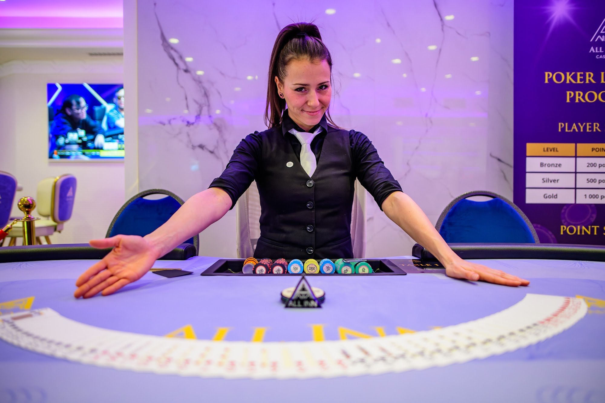 A young woman with dark hair in a high ponytail and wearing a black casino dealer outfit with a white tie, smiling and standing behind a poker table with poker chips and a dealer button, in a casino gaming area with purple lighting and wall decor.