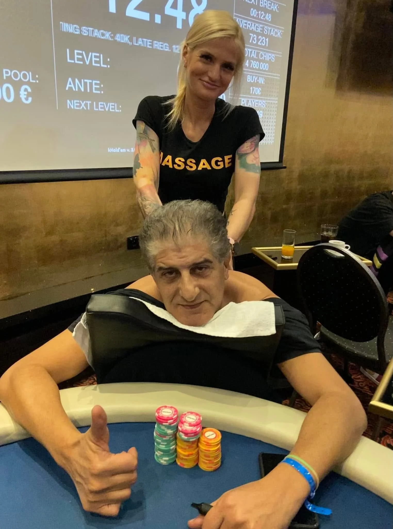 A woman with blonde hair and tattoos on her arms smiling and working at a poker table, with a stack of poker chips in front of her, and a man sitting at the table with his head down. There is a large screen in the background displaying numbers and in