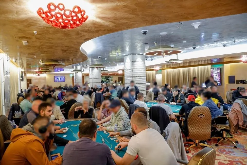 People playing poker at a crowded casino poker room with green tables, gold curtains, and decorative ceiling lights.