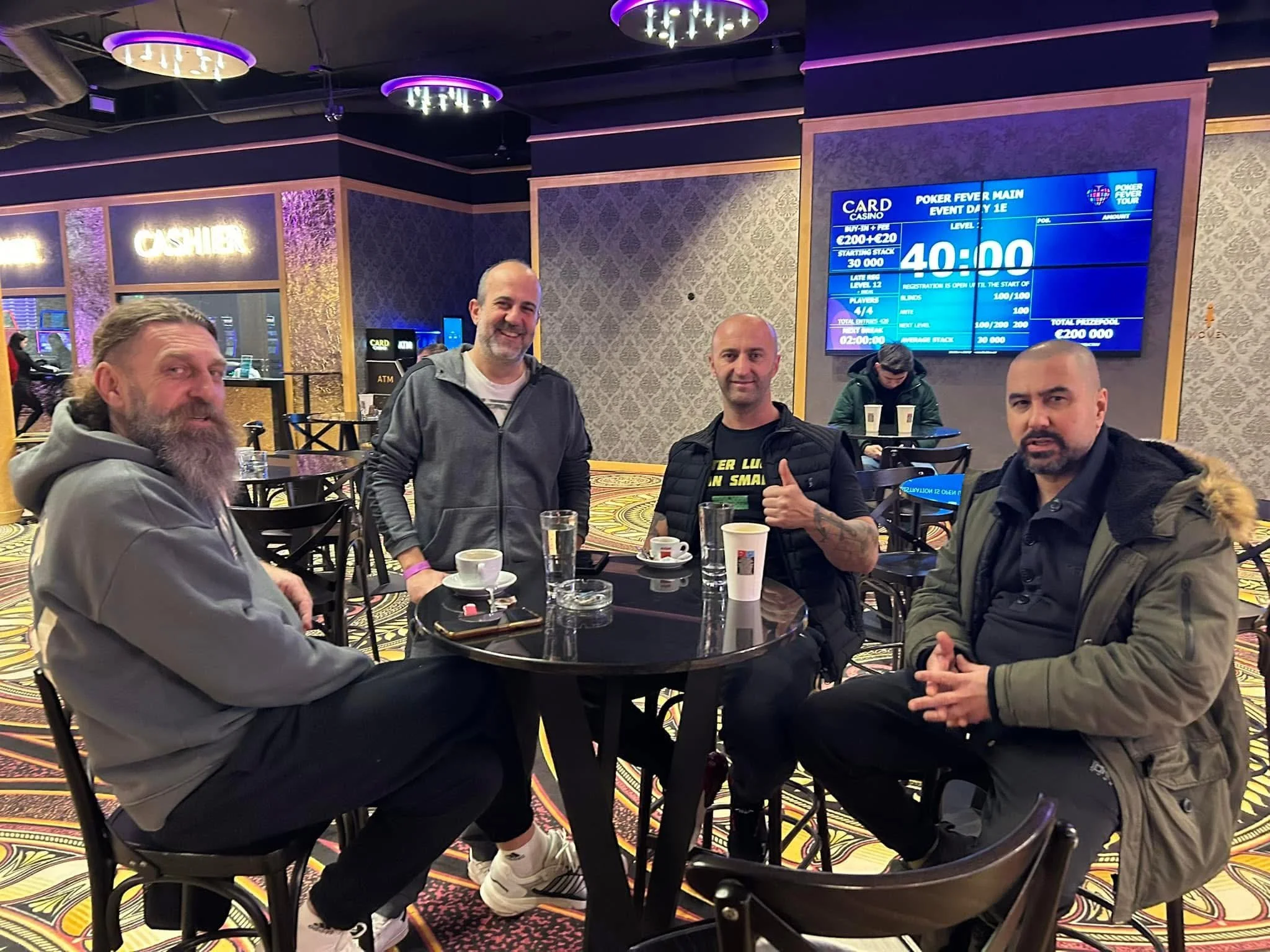 Four men sitting around a round table at a casino, with cups and glasses on the table, smiling and looking at the camera. A large digital screen displays poker tournament information in the background.