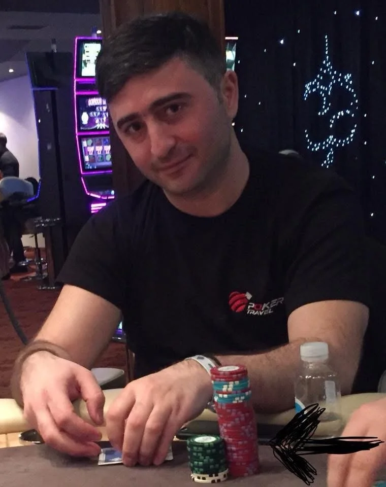 A man sitting at a poker table in a casino, handling poker chips with a slight smile. There are slot machines and decorative lights in the background.
