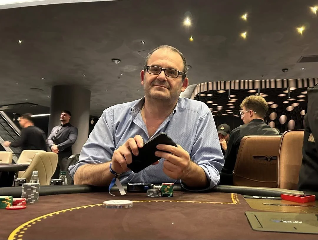 A man with glasses and a blue shirt sitting at a casino table with poker chips, holding a smartphone.
