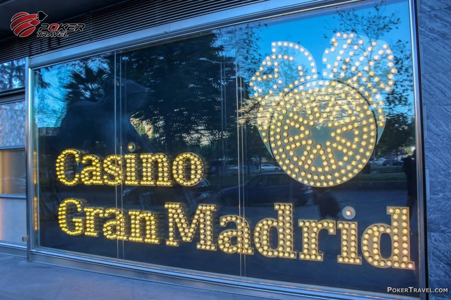 Large illuminated sign reading "Casino Gran Madrid" with a roulette wheel graphic on a glass window, reflecting trees and the street outside.
