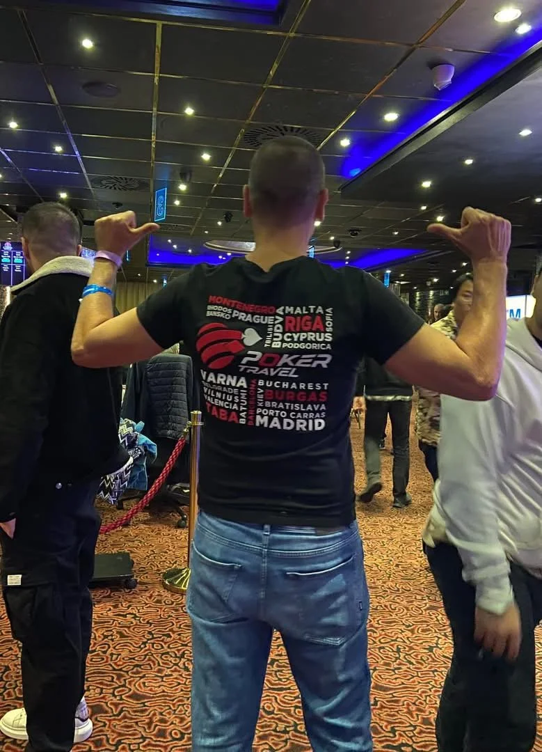 A man with a shaved head flexing his biceps at an indoor event, wearing a black T-shirt with a map and city names of European countries, and jeans. Other people are around.