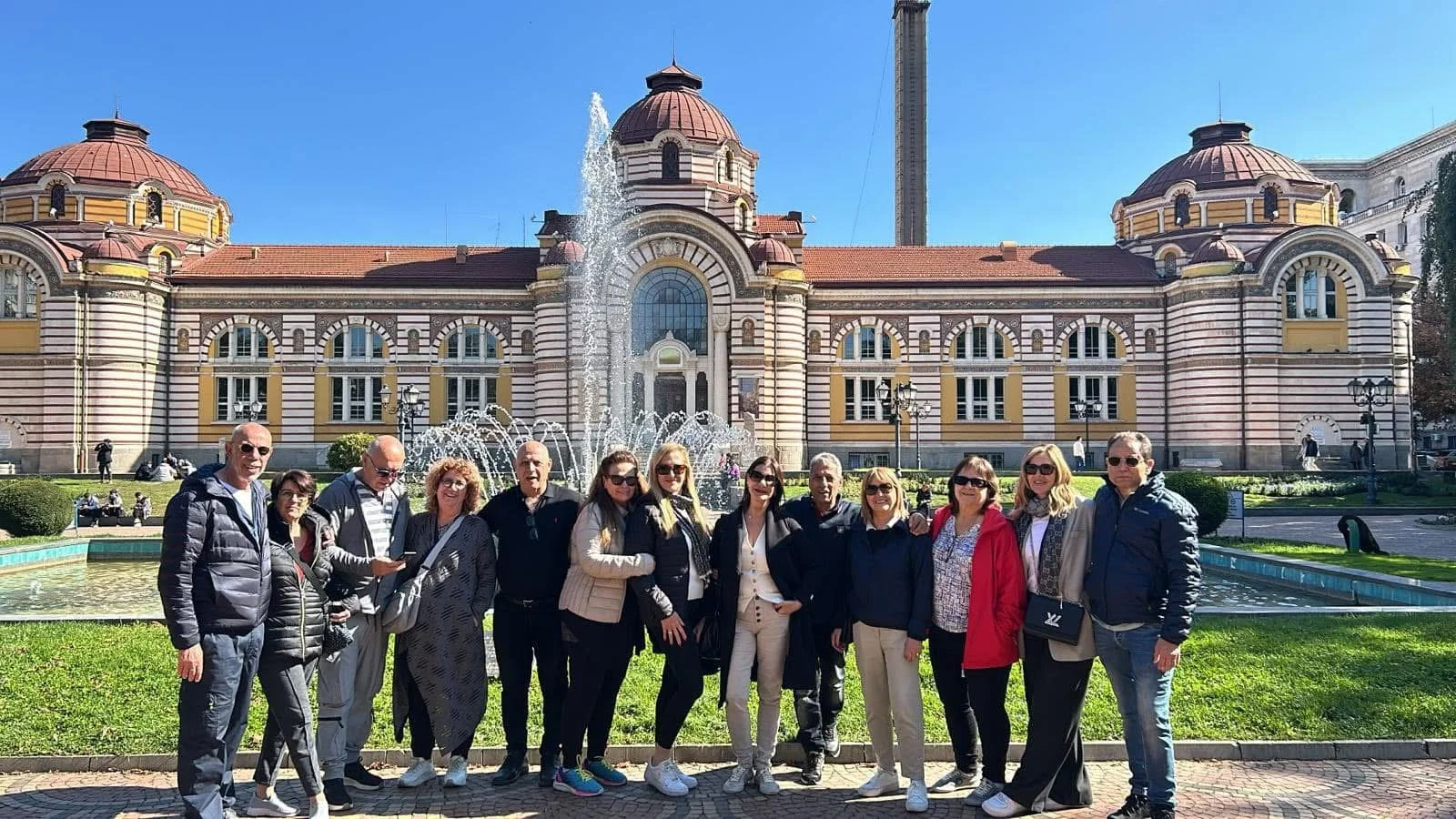 Group of people posing in front of a historic building with fountains, under a clear blue sky.