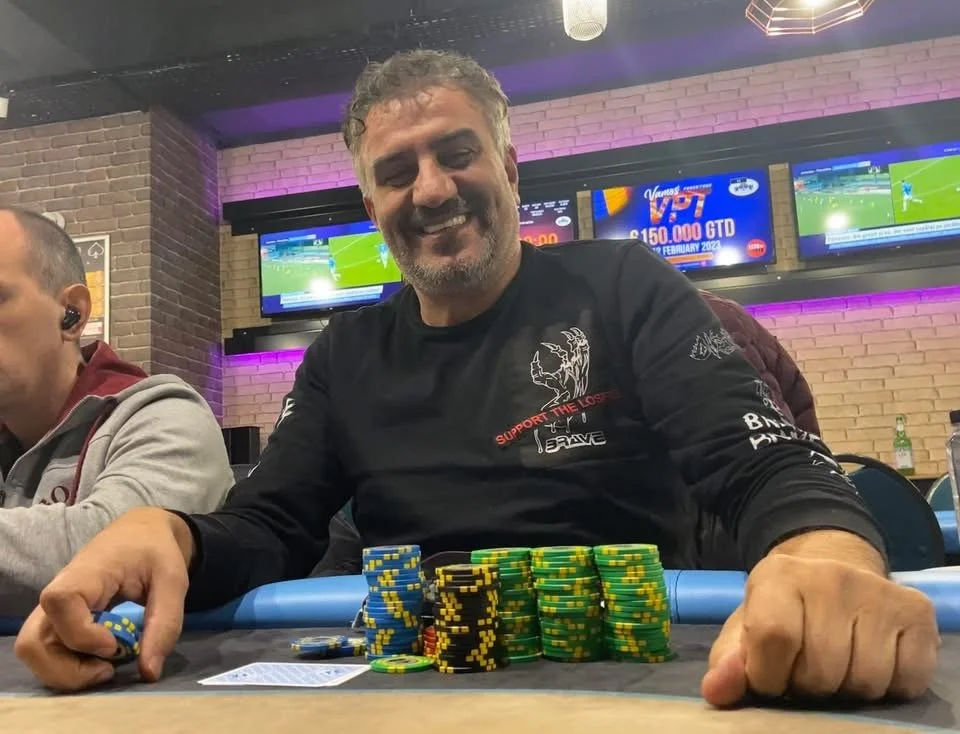 Man smiling at poker table with colorful poker chips, in a casino or gaming setting, with multiple screens in background showing sports and betting information.