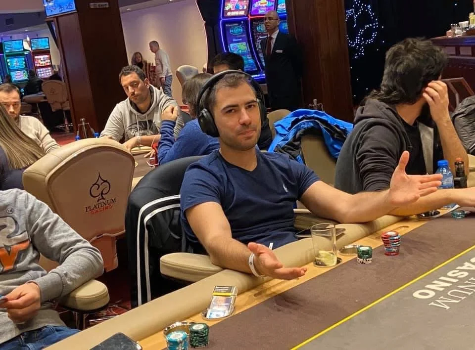 Man sitting at a poker table with headphones, gesturing with both hands, with poker chips and a drink in front of him, at a casino.