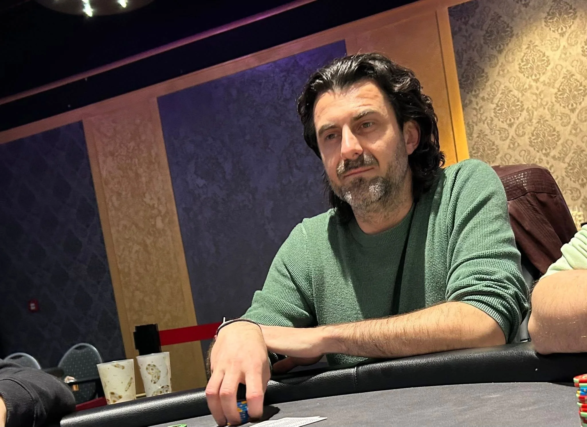 A man with dark hair and a beard is sitting at a poker table. He is wearing a green sweater and appears to be focused on the game. The background features textured purple and gold wallpaper.