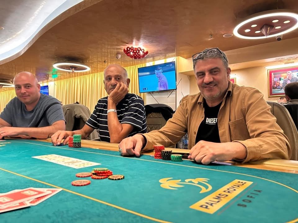 Three men playing poker at a casino table with poker chips, in a room with yellow curtains and a television screen in the background.