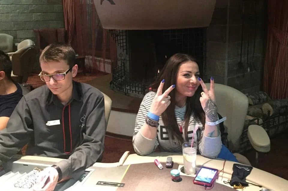 Woman with long dark hair making peace signs near her eyes, sitting at a table in a restaurant or casino, with a drink and mobile phone in front of her, and a man with glasses and a gray shirt sitting next to her.