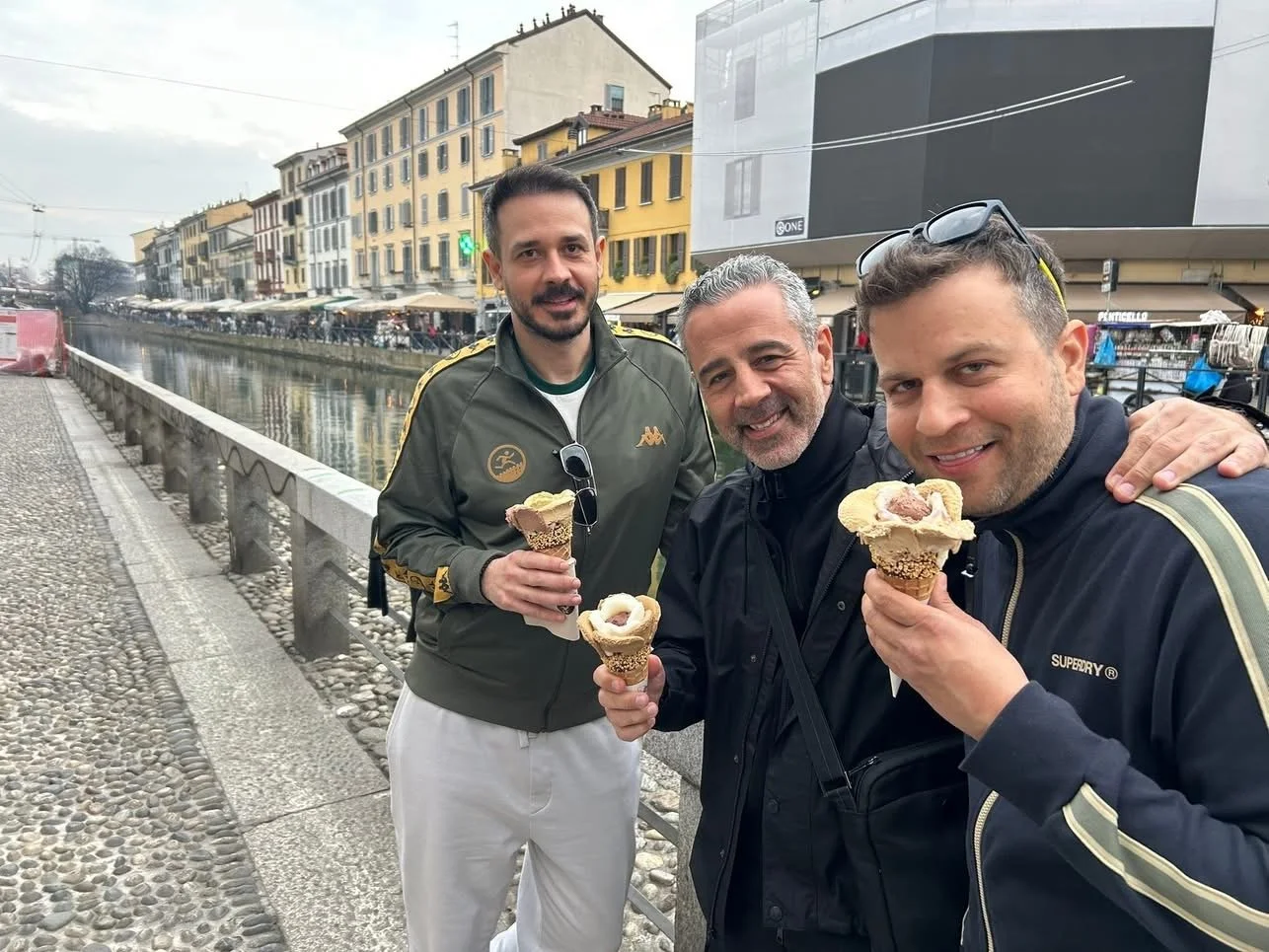 Three men standing by a canal, holding ice cream cones and smiling, with a city street and buildings in the background.
