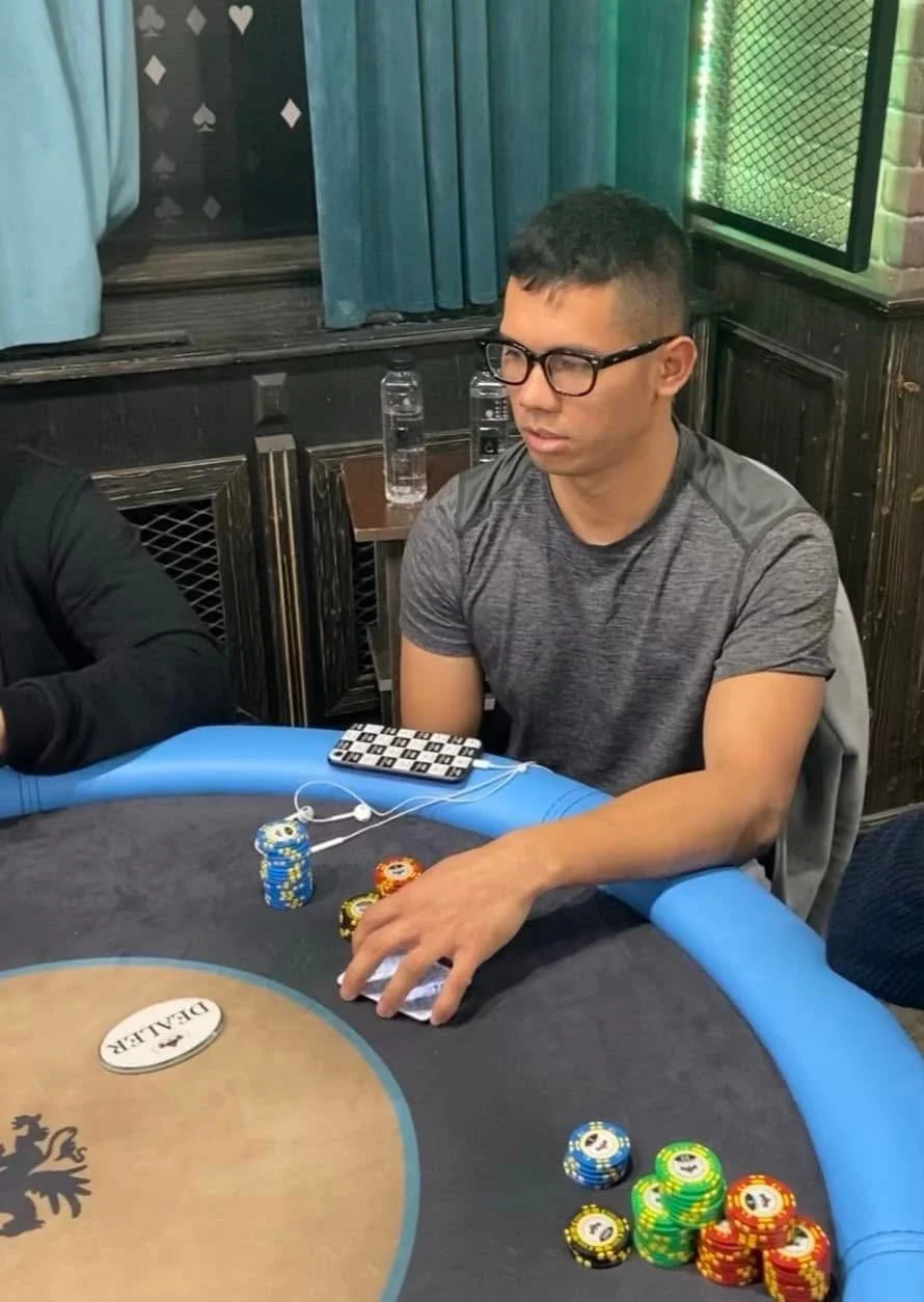 A young man with glasses and a gray t-shirt sitting at a poker table with chips and a deck of cards, in a dimly lit room with dark wooden walls and a green netted window.