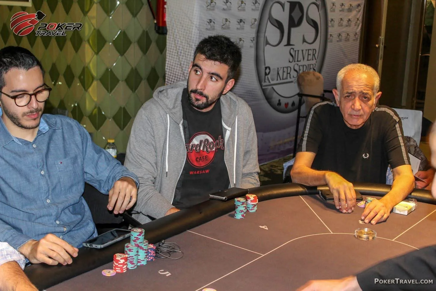 Three men sitting around a poker table with chips, awards, and a PokerTravel logo in the background.