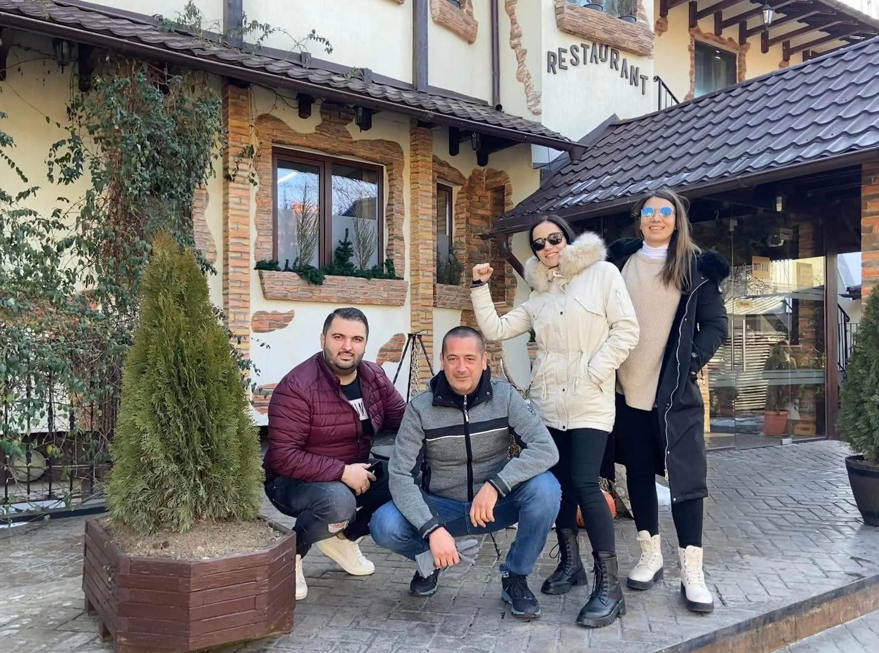 Group of five people posing in front of a restaurant building with decorative brickwork and windows. Two men are squatting in front, and three women are standing behind, wearing casual winter clothing and sunglasses.