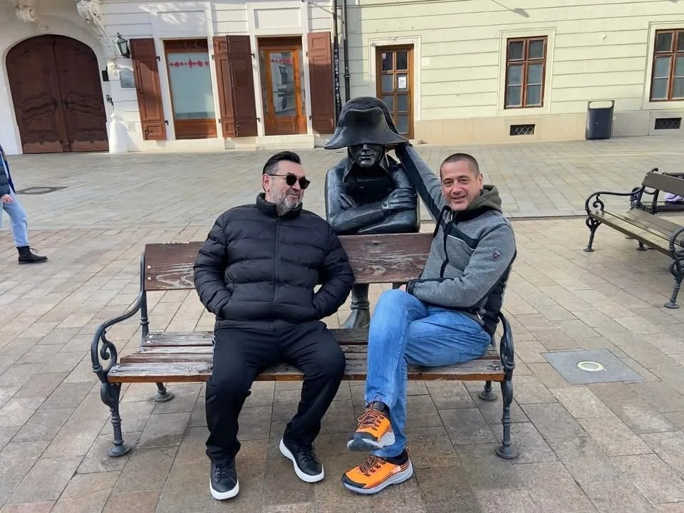 Two men sitting on a wooden bench next to a sculpture of a person wearing a hat. One man is wearing a black jacket and black pants, the other is wearing a gray hooded jacket and blue jeans. They are smiling and appear to be enjoying their time outsid