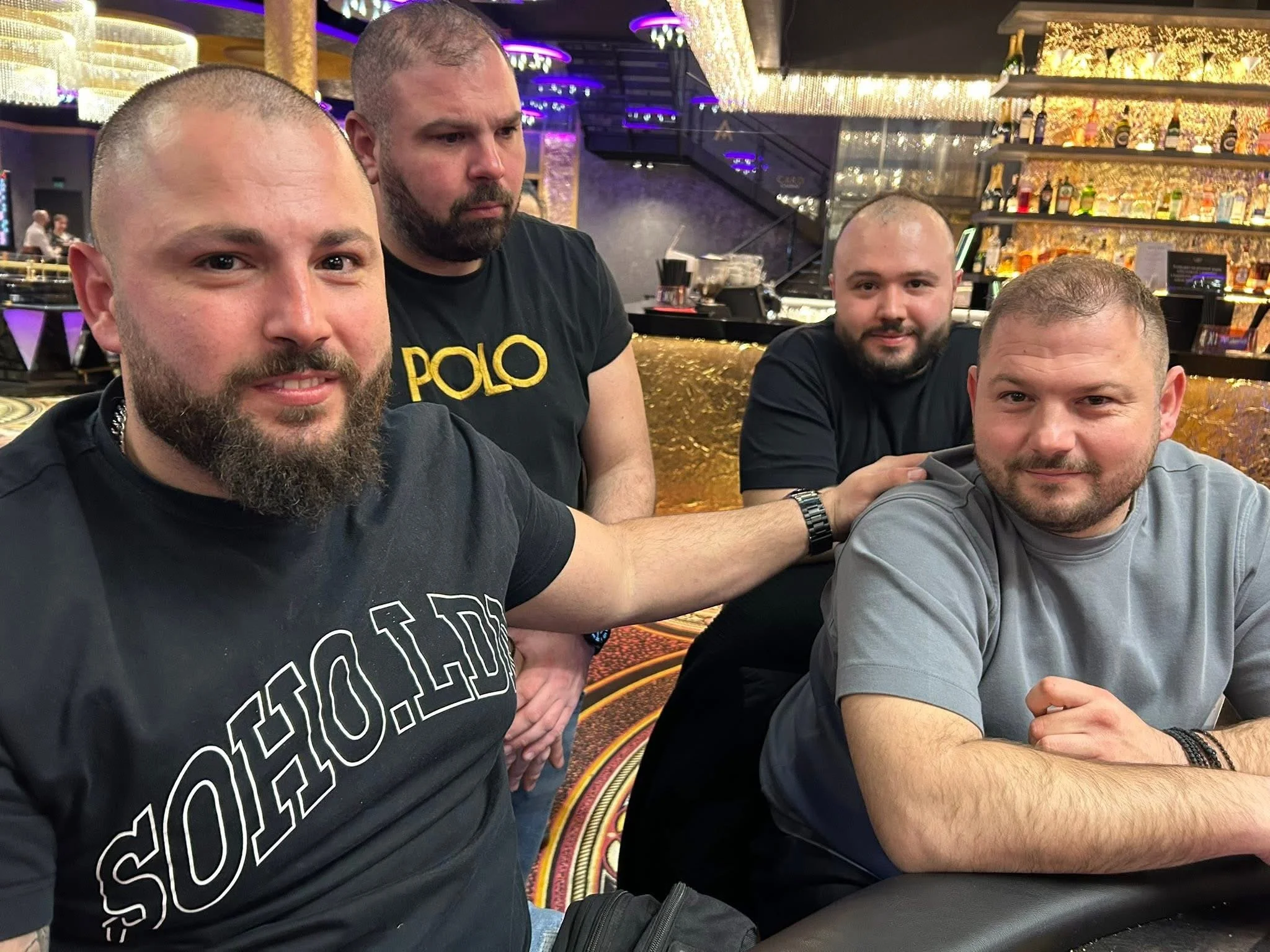 Four men sitting at a bar in a casino. They are smiling and looking at the camera. The background has shelves with bottles and colorful lights.