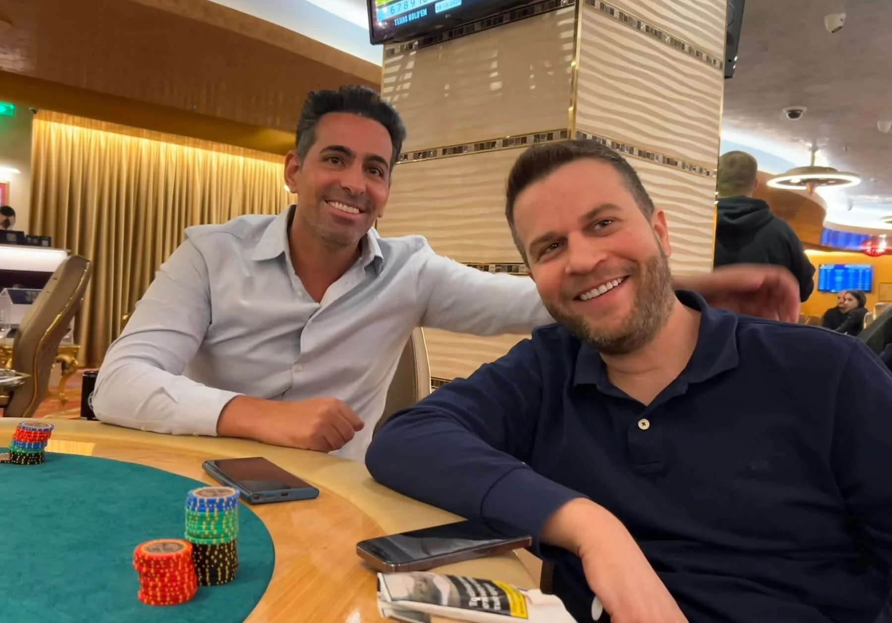Two men smiling at a poker table in a casino, with poker chips, a phone, and a magazine on the table. One man is wearing a white shirt, the other a dark shirt, and there are other people and digital screens in the background.