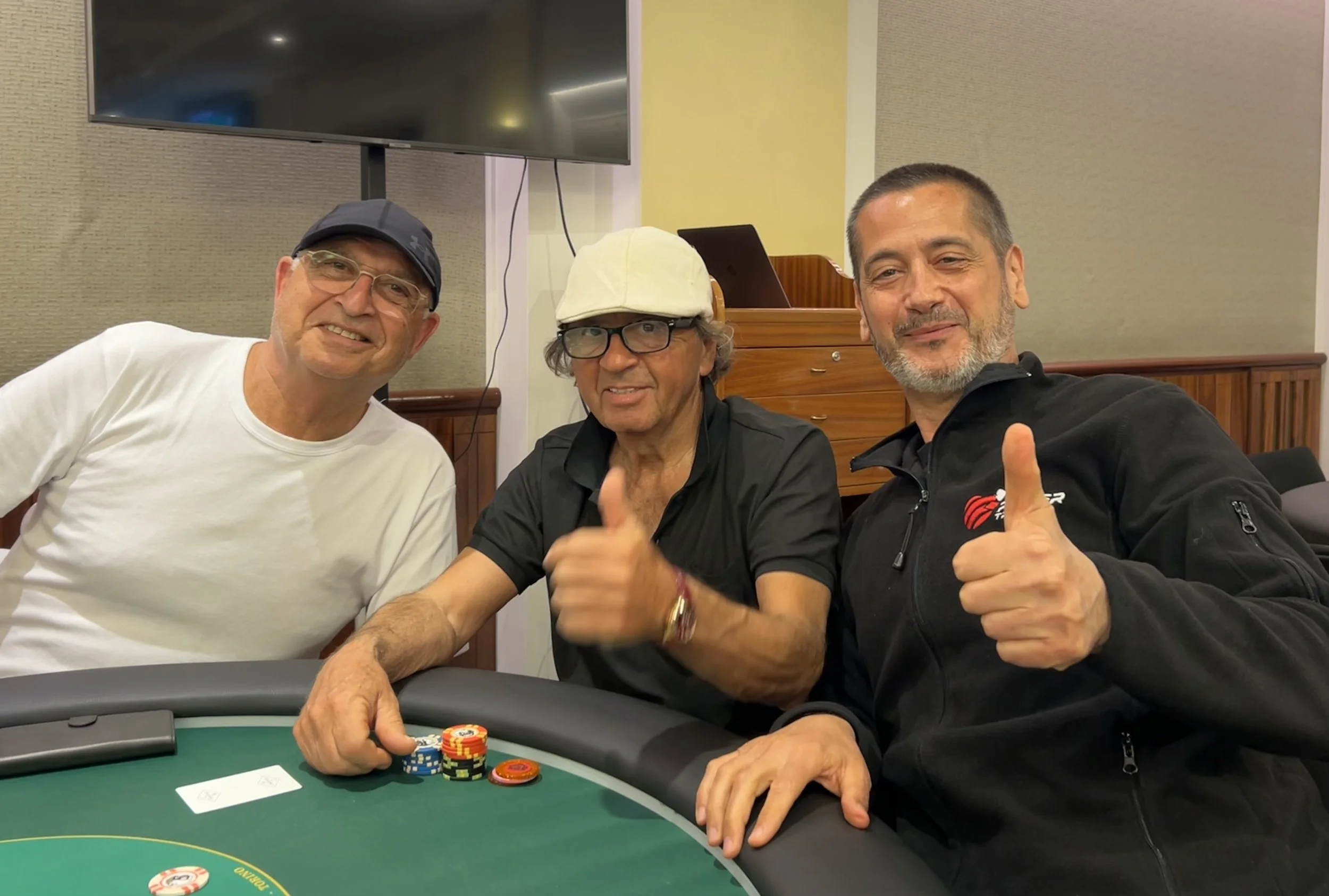 Three men sitting at a poker table with stacks of chips, smiling and giving thumbs up, in a room with a large television and wood paneling.
