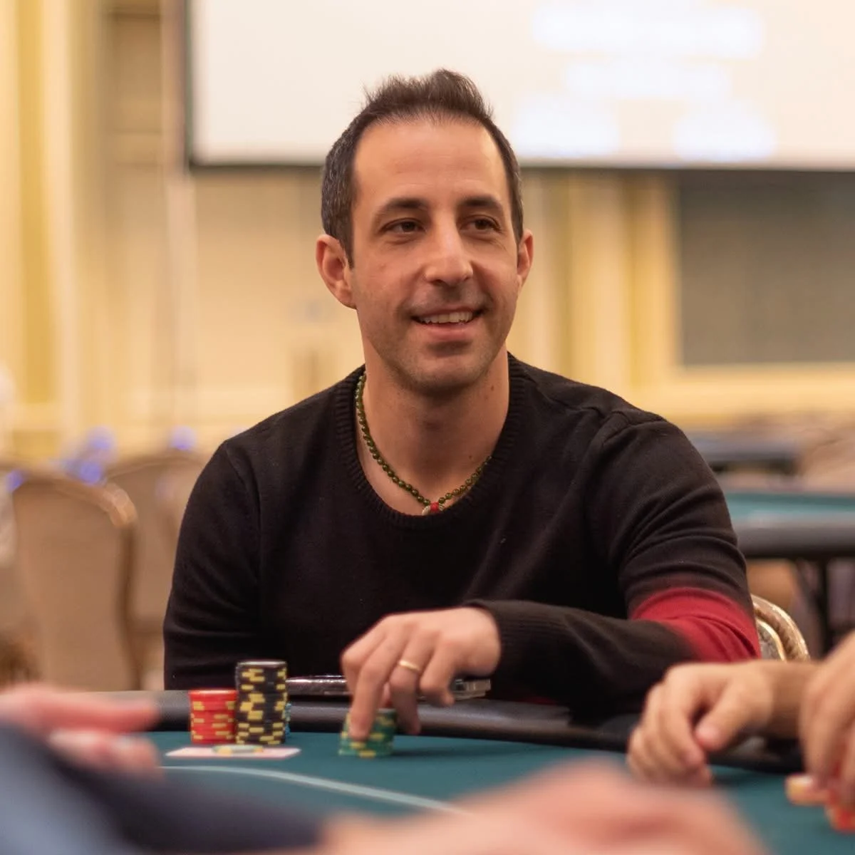A man smiling at a poker table with poker chips in front of him, in a well-lit room with yellow walls and a large screen in the background.